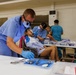 A field or community dental clinic is in operation inside a large indoor hall or gymnasium. Multiple dental practitioners in scrubs and protective gear are simultaneously treating patients seated in portable chairs. The setting appears to be a temporary or mobile dental outreach event with supplies organized on folding tables.