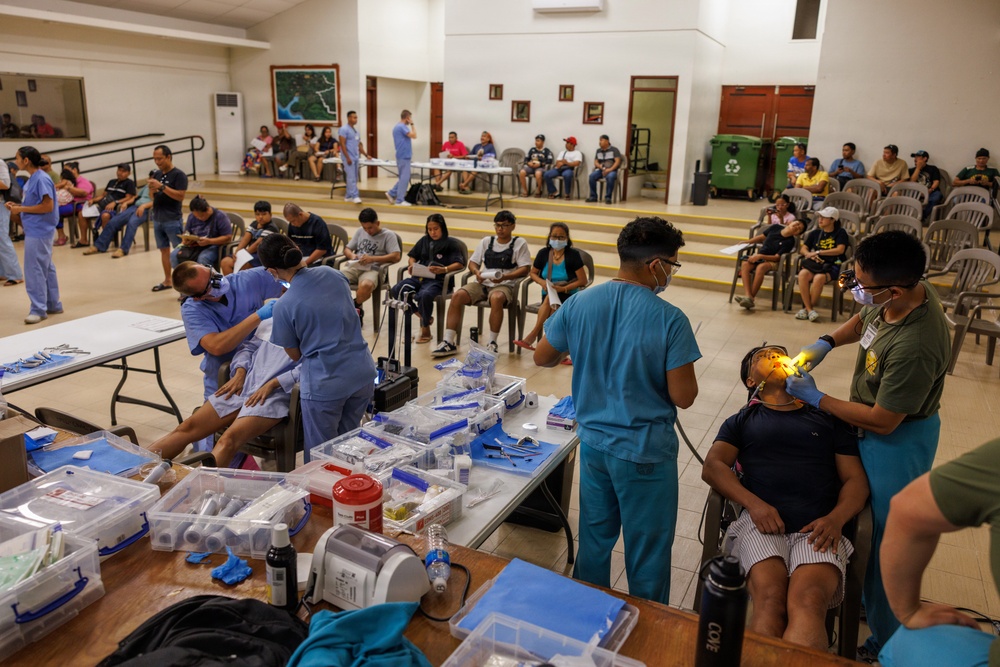 A large-scale community medical/dental outreach event taking place in what appears to be a community hall or gymnasium. Multiple healthcare providers in scrubs are simultaneously treating patients, while a large number of community members wait in chairs arranged throughout the room. Dental procedures appear to be the primary service being provided, with portable equipment set up on folding tables.
