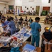 A large-scale community medical/dental outreach event taking place in what appears to be a community hall or gymnasium. Multiple healthcare providers in scrubs are simultaneously treating patients, while a large number of community members wait in chairs arranged throughout the room. Dental procedures appear to be the primary service being provided, with portable equipment set up on folding tables.