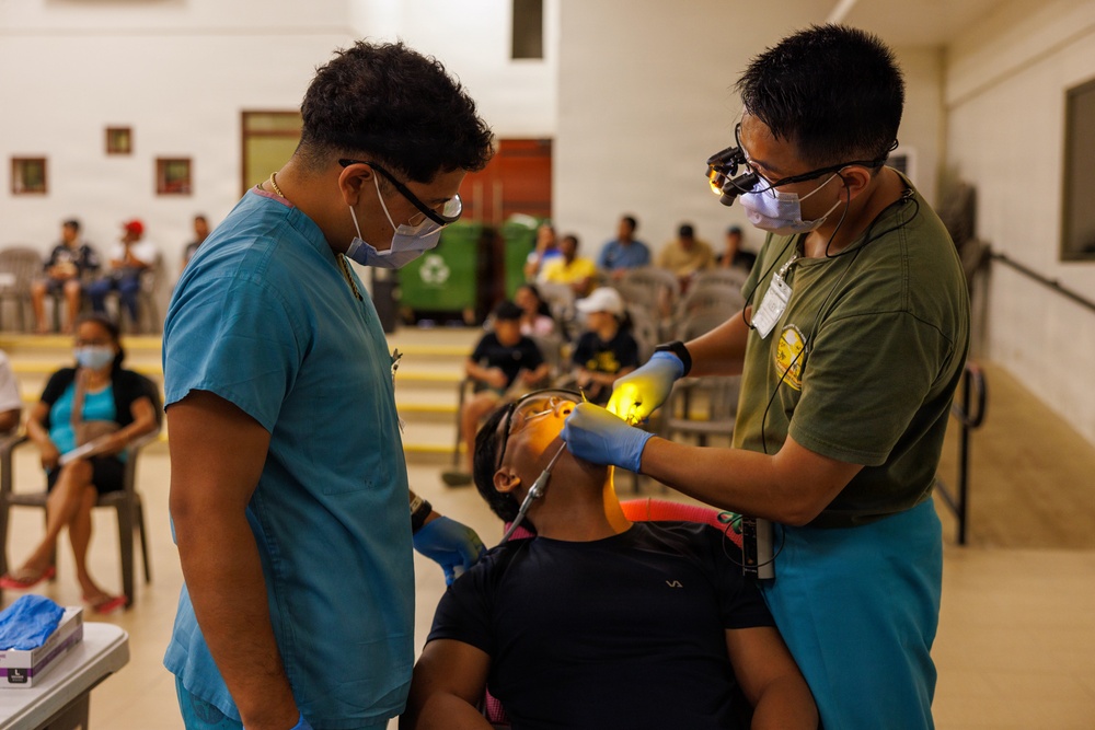 A dental procedure is being performed on a seated patient in what appears to be a community health outreach setting. Two medical personnel wearing scrubs, masks, and protective eyewear are treating the patient, with one using a dental instrument and a light source. A waiting area with multiple people seated in chairs is visible in the background.