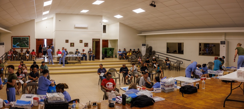 A large community hall has been converted into a temporary medical clinic or health screening event. Multiple healthcare workers in blue scrubs are attending to numerous civilians seated in chairs throughout the space. The setting appears to be an organized medical outreach or humanitarian assistance event.