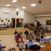 A large community hall has been converted into a temporary medical clinic or health screening event. Multiple healthcare workers in blue scrubs are attending to numerous civilians seated in chairs throughout the space. The setting appears to be an organized medical outreach or humanitarian assistance event.