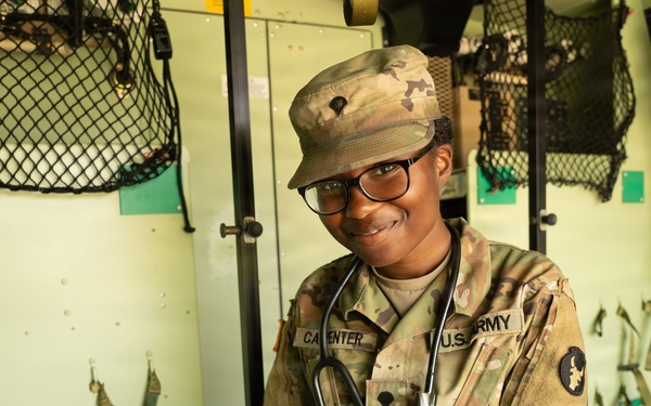 A U.S. Army soldier with the last name 'Carpenter' is seated inside what appears to be a military medical vehicle or ambulance. The soldier is wearing OCP (Operational Camouflage Pattern) uniform with a stethoscope around their neck, indicating a medical role. They are smiling at the camera in a relaxed, posed manner.