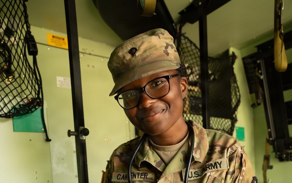 A U.S. Army soldier named Carpenter, wearing OCP (Operational Camouflage Pattern) uniform with a stethoscope around their neck, is photographed inside what appears to be a military medical vehicle or aid station. The soldier is smiling at the camera while organizing or inspecting a military medical kit/aid bag. The setting and equipment indicate this is a combat medic or medical specialist.