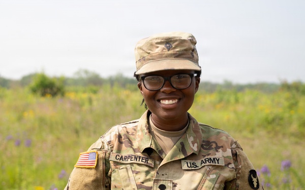 A U.S. Army soldier named Carpenter stands smiling in an outdoor field setting wearing full OCP (Operational Camouflage Pattern) uniform. The soldier is posed with hands behind their back in a relaxed, confident stance. The background features a lush meadow with wildflowers under an overcast sky.