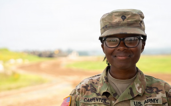 A U.S. Army soldier named Carpenter is photographed in a portrait-style image wearing Operational Camouflage Pattern (OCP) uniform. The soldier is smiling and standing with hands on hips against a blurred outdoor background that appears to be a military training or base area. The image appears to be an official or semi-official military portrait.