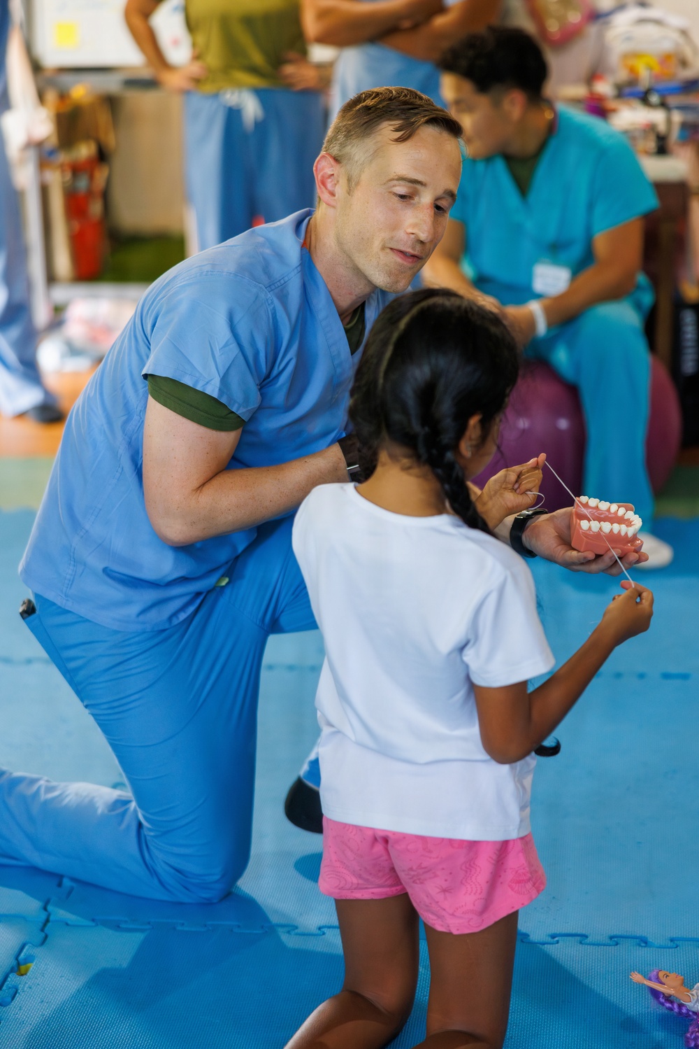 A dental healthcare professional in blue scrubs kneels down to teach a young girl how to floss using a large dental model. The setting appears to be a community health outreach or educational event, with other medical staff visible in the background. The interaction is hands-on and child-focused, demonstrating proper oral hygiene technique.