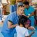 A dental healthcare professional in blue scrubs kneels down to teach a young girl how to floss using a large dental model. The setting appears to be a community health outreach or educational event, with other medical staff visible in the background. The interaction is hands-on and child-focused, demonstrating proper oral hygiene technique.