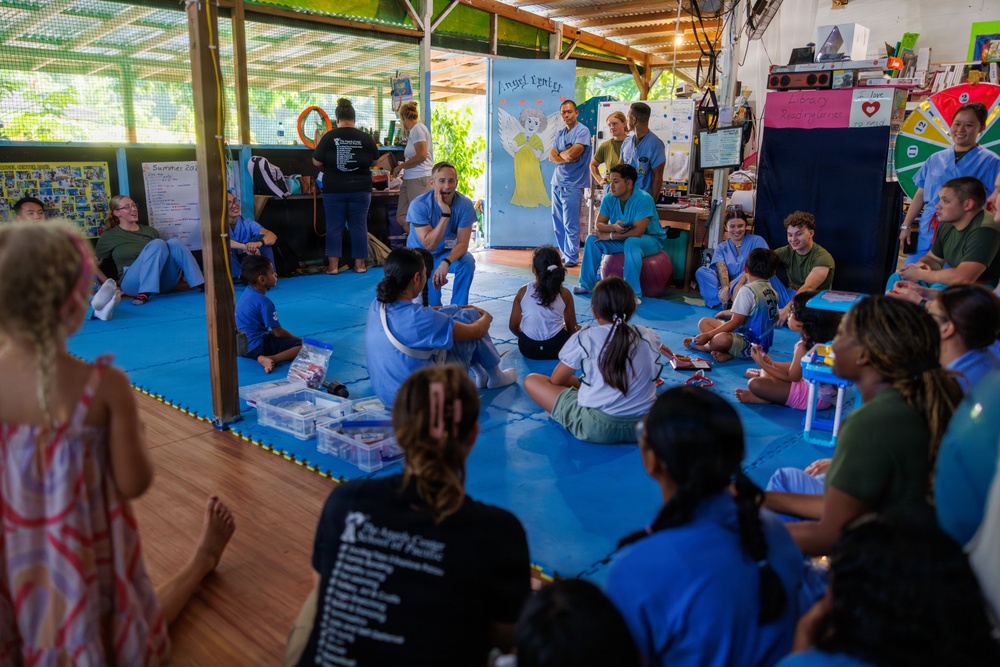 A group of medical personnel wearing blue scrubs are gathered with children and adults on a blue mat floor inside what appears to be a community center or educational facility. The setting suggests a medical outreach or volunteer health mission, with participants sitting in an informal circle arrangement. A painted mural reading 'Angel Center' is visible on a white board in the background, indicating this may be a charitable or faith-based organization.