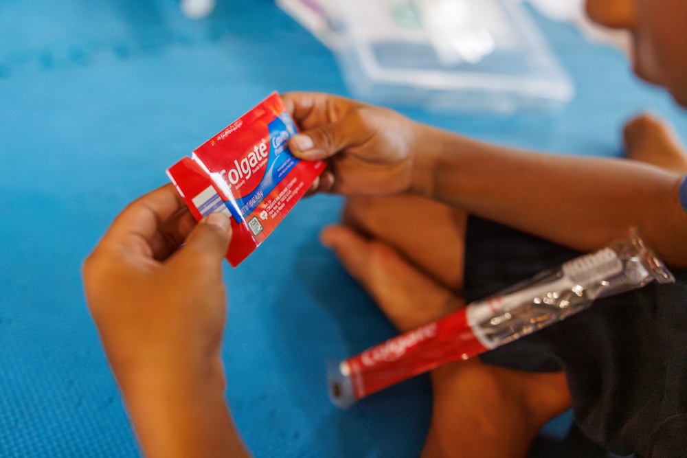 A child is holding a red Colgate toothpaste tube with both hands, examining it closely. A Colgate toothbrush in clear packaging is also visible resting on the child's lap. The setting appears to be on a blue foam mat or floor surface.