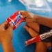 A child is holding a red Colgate toothpaste tube with both hands, examining it closely. A Colgate toothbrush in clear packaging is also visible resting on the child's lap. The setting appears to be on a blue foam mat or floor surface.