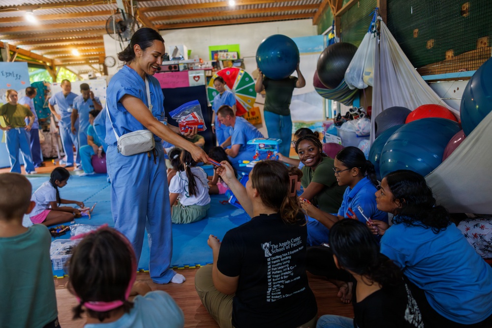 A group of medical personnel in blue scrubs are engaged in a community outreach or volunteer event, interacting with children and adults in what appears to be an open-air facility or community center. A smiling healthcare worker in blue scrubs is distributing items, likely supplies or small gifts, to seated participants. The setting includes colorful exercise balls, educational materials, and a lively, informal atmosphere suggesting a health or wellness outreach program.
