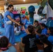 A group of medical personnel in blue scrubs are engaged in a community outreach or volunteer event, interacting with children and adults in what appears to be an open-air facility or community center. A smiling healthcare worker in blue scrubs is distributing items, likely supplies or small gifts, to seated participants. The setting includes colorful exercise balls, educational materials, and a lively, informal atmosphere suggesting a health or wellness outreach program.