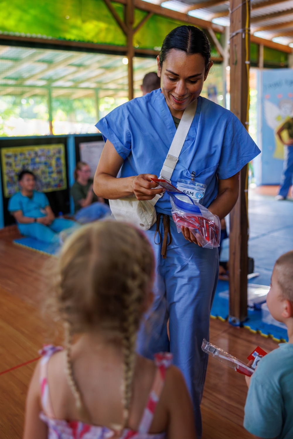 A healthcare worker named 'SAM,' wearing blue scrubs and a name badge, is smiling while distributing items from a clear plastic bag to young children in an open-air pavilion setting. The scene appears to be a community health outreach or dental hygiene event, with other personnel visible in the background. The items being distributed appear to be red-handled toothbrushes in individual packaging.