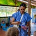 A healthcare worker named 'SAM,' wearing blue scrubs and a name badge, is smiling while distributing items from a clear plastic bag to young children in an open-air pavilion setting. The scene appears to be a community health outreach or dental hygiene event, with other personnel visible in the background. The items being distributed appear to be red-handled toothbrushes in individual packaging.