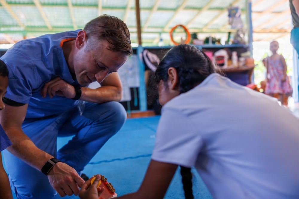 A person wearing blue medical scrubs and a fitness tracker is crouching down and interacting with a child or young person, appearing to demonstrate or show a dental model (teeth model) to them. The setting appears to be an informal community space or open-air building with blue floor mats. This appears to be a medical or dental outreach or educational activity in what looks like a developing-world community setting.