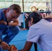 A person wearing blue medical scrubs and a fitness tracker is crouching down and interacting with a child or young person, appearing to demonstrate or show a dental model (teeth model) to them. The setting appears to be an informal community space or open-air building with blue floor mats. This appears to be a medical or dental outreach or educational activity in what looks like a developing-world community setting.