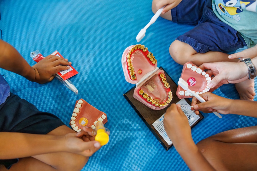 Children are engaged in a dental hygiene education activity, using oversized tooth models and toothbrushes to learn proper brushing techniques. The scene appears to be a classroom or educational setting with children sitting on a blue foam mat. Two dental models are visible — one showing healthy white teeth and another showing decayed/discolored teeth, likely used for comparison purposes.