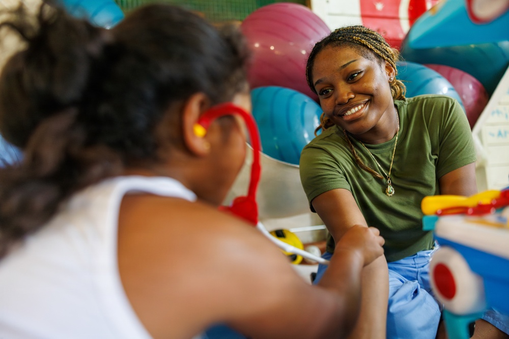 A smiling young adult woman in a green t-shirt interacts warmly with a young child in what appears to be a play or therapy room. The child, seen from behind, is wearing red-framed glasses and a white top. Colorful exercise balls and toys are visible in the background.