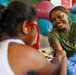 A smiling young adult woman in a green t-shirt interacts warmly with a young child in what appears to be a play or therapy room. The child, seen from behind, is wearing red-framed glasses and a white top. Colorful exercise balls and toys are visible in the background.