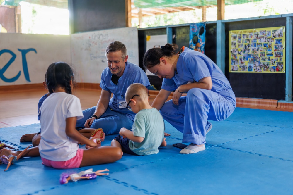 Two healthcare workers in blue scrubs are seated on a blue foam mat interacting with two young children in what appears to be a community center or school setting. The male worker, whose name tag reads 'CODY,' is smiling warmly at the children while the female worker leans in attentively. The scene suggests a medical outreach or volunteer mission involving child engagement.