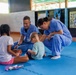 Two healthcare workers in blue scrubs are seated on a blue foam mat interacting with two young children in what appears to be a community center or school setting. The male worker, whose name tag reads 'CODY,' is smiling warmly at the children while the female worker leans in attentively. The scene suggests a medical outreach or volunteer mission involving child engagement.