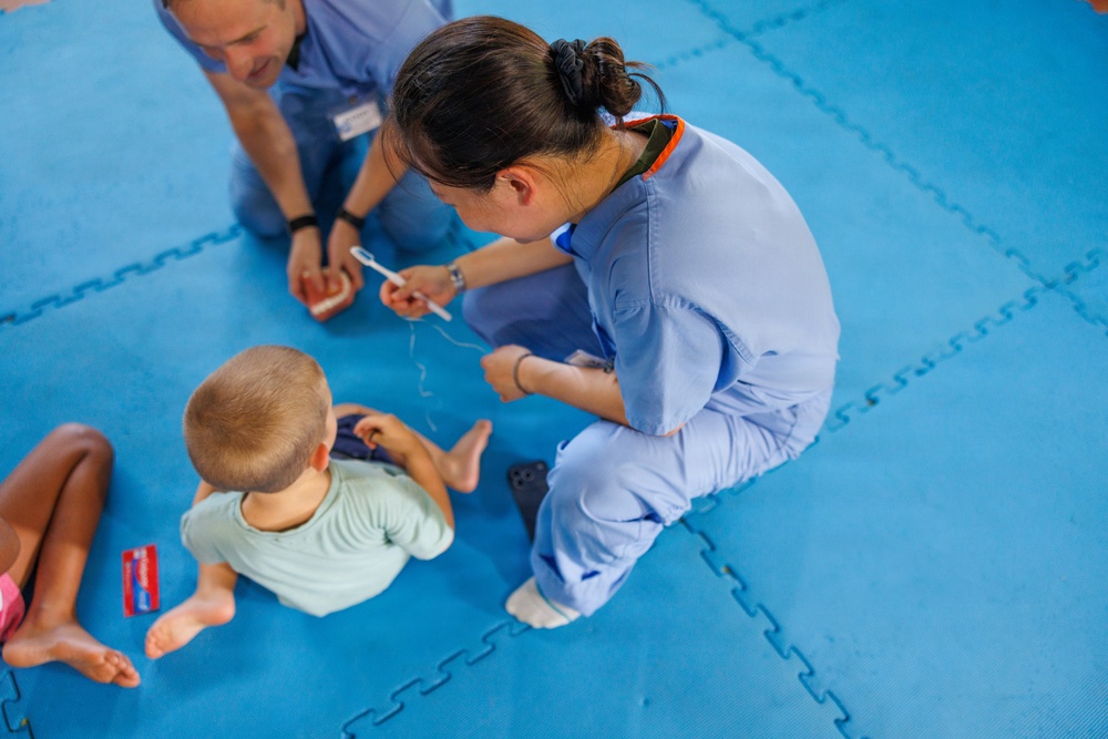 Two healthcare workers in blue scrubs are seated on a blue foam puzzle mat, interacting with at least two young children. One worker appears to be demonstrating toothbrushing technique, holding a toothbrush toward a baby/toddler in a light green shirt. A red toothpaste box (appears to be Colgate) is visible on the mat, suggesting a dental hygiene education session.