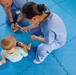 Two healthcare workers in blue scrubs are seated on a blue foam puzzle mat, interacting with at least two young children. One worker appears to be demonstrating toothbrushing technique, holding a toothbrush toward a baby/toddler in a light green shirt. A red toothpaste box (appears to be Colgate) is visible on the mat, suggesting a dental hygiene education session.