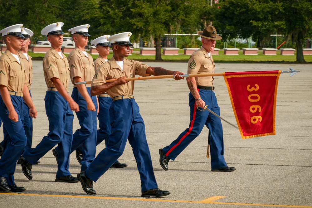 A group of United States Marine Corps recruits or soldiers are marching in formation during what appears to be a graduation or parade ceremony. A drill instructor wearing a campaign cover (drill sergeant hat) leads the formation while carrying a guidon flag marked '3066'. The scene takes place on a paved parade deck with green trees visible in the background.