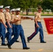 A group of United States Marine Corps recruits or soldiers are marching in formation during what appears to be a graduation or parade ceremony. A drill instructor wearing a campaign cover (drill sergeant hat) leads the formation while carrying a guidon flag marked '3066'. The scene takes place on a paved parade deck with green trees visible in the background.