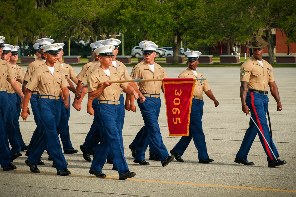 A formation of United States Marine Corps recruits or graduates is marching in a parade or graduation ceremony. They are carrying a red guidon pennant bearing the number '3065', indicating Platoon 3065. A drill instructor wearing a distinctive campaign cover (Smokey Bear hat) leads the formation from the right side.