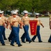 A formation of United States Marine Corps recruits or graduates is marching in a parade or graduation ceremony. They are carrying a red guidon pennant bearing the number '3065', indicating Platoon 3065. A drill instructor wearing a distinctive campaign cover (Smokey Bear hat) leads the formation from the right side.