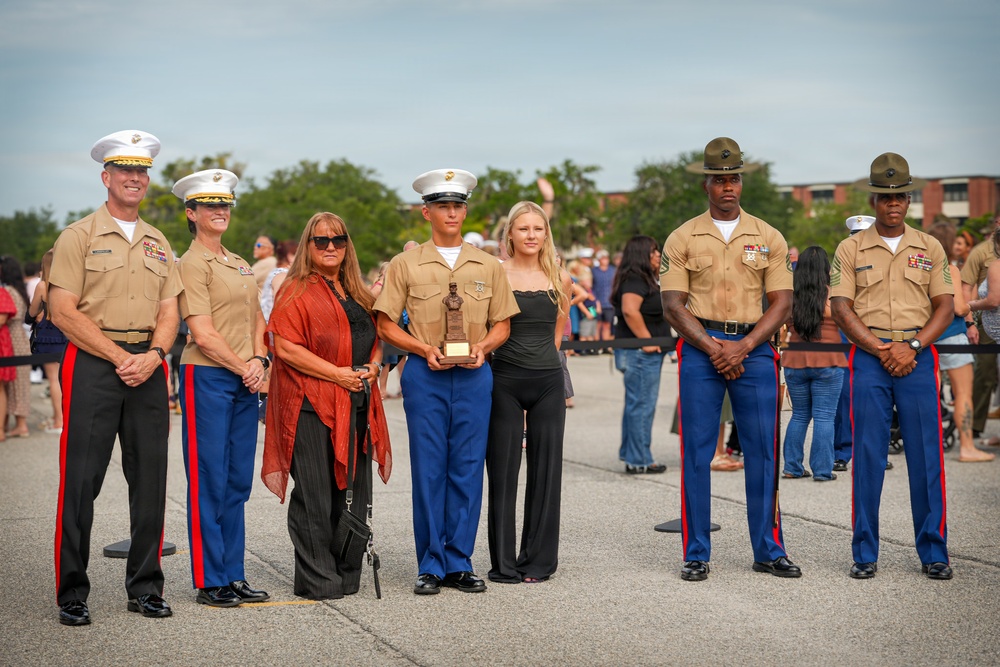 A formal U.S. Marine Corps ceremony is taking place outdoors, likely a graduation or awards event. A young Marine in the center is holding a trophy or award, flanked by senior officers and what appear to be family members. The group is posed for a commemorative photograph on a paved parade deck.