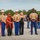 A formal U.S. Marine Corps ceremony is taking place outdoors, likely a graduation or awards event. A young Marine in the center is holding a trophy or award, flanked by senior officers and what appear to be family members. The group is posed for a commemorative photograph on a paved parade deck.