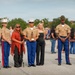 A formal U.S. Marine Corps ceremony is taking place outdoors, likely a graduation or awards event. A young Marine in the center is holding a trophy or award, flanked by senior officers and what appear to be family members. The group is posed for a commemorative photograph on a paved parade deck.