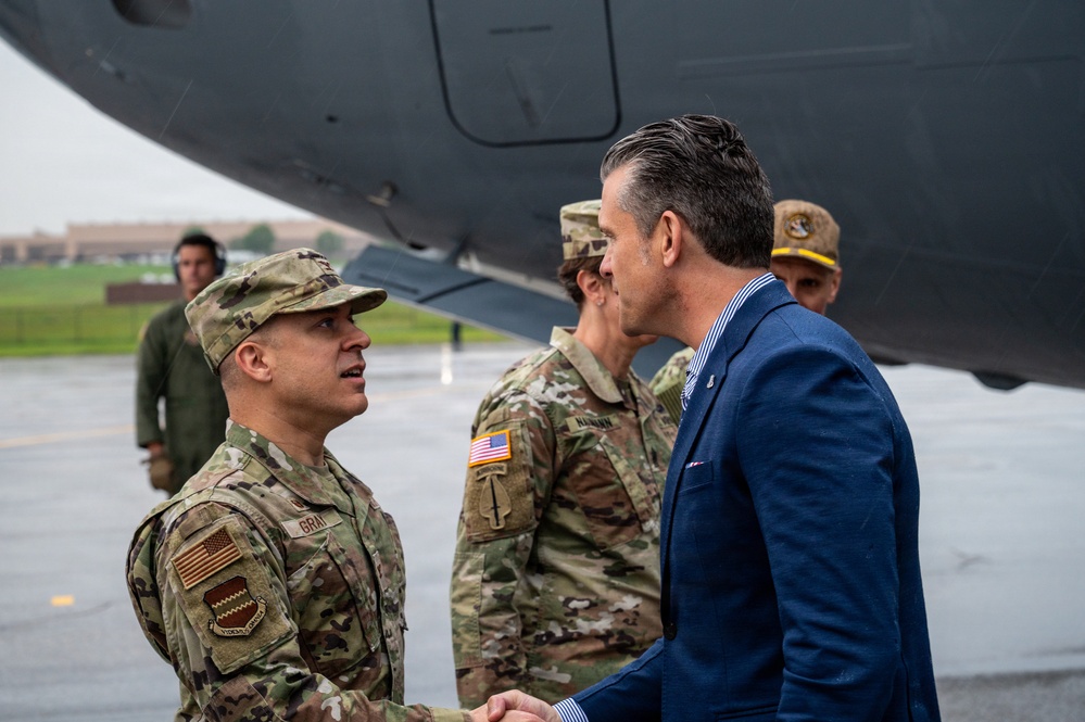 A military greeting ceremony is taking place on an airfield tarmac, likely at a U.S. Air Force base. A uniformed soldier with the name tape 'GRAY' and U.S. Army/Air Force patches is shaking hands with a civilian man in a blue suit. Several other uniformed personnel are visible in the background near what appears to be a large military aircraft.