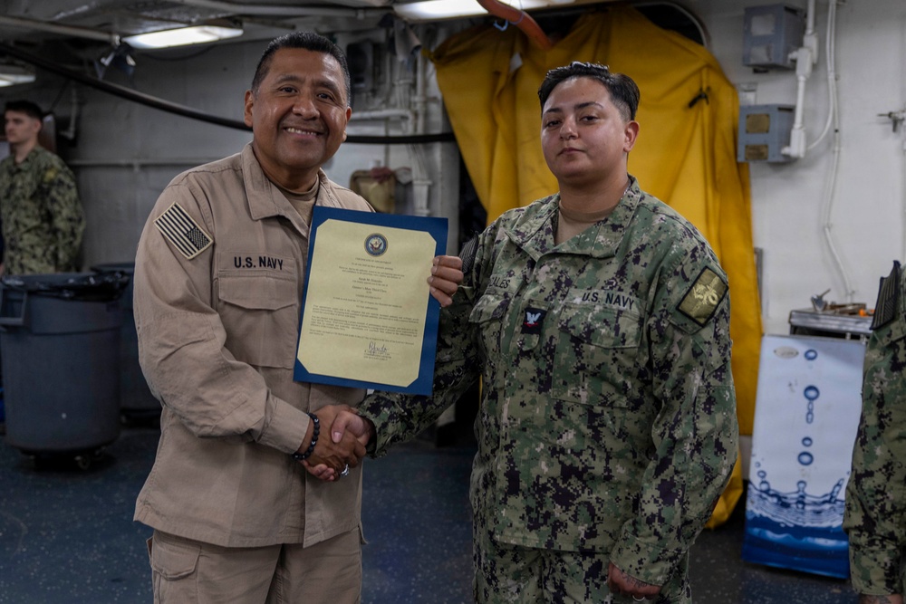 Two U.S. Navy personnel are posing for a photo during what appears to be a formal recognition or award ceremony aboard a naval vessel. The individual on the left, wearing tan/khaki Navy working uniform, is presenting a certificate to the individual on the right, who is wearing the Navy Working Uniform (NWU) digital camouflage pattern. Both are shaking hands while jointly holding the framed certificate.