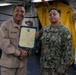 Two U.S. Navy personnel are posing for a photo during what appears to be a formal recognition or award ceremony aboard a naval vessel. The individual on the left, wearing tan/khaki Navy working uniform, is presenting a certificate to the individual on the right, who is wearing the Navy Working Uniform (NWU) digital camouflage pattern. Both are shaking hands while jointly holding the framed certificate.