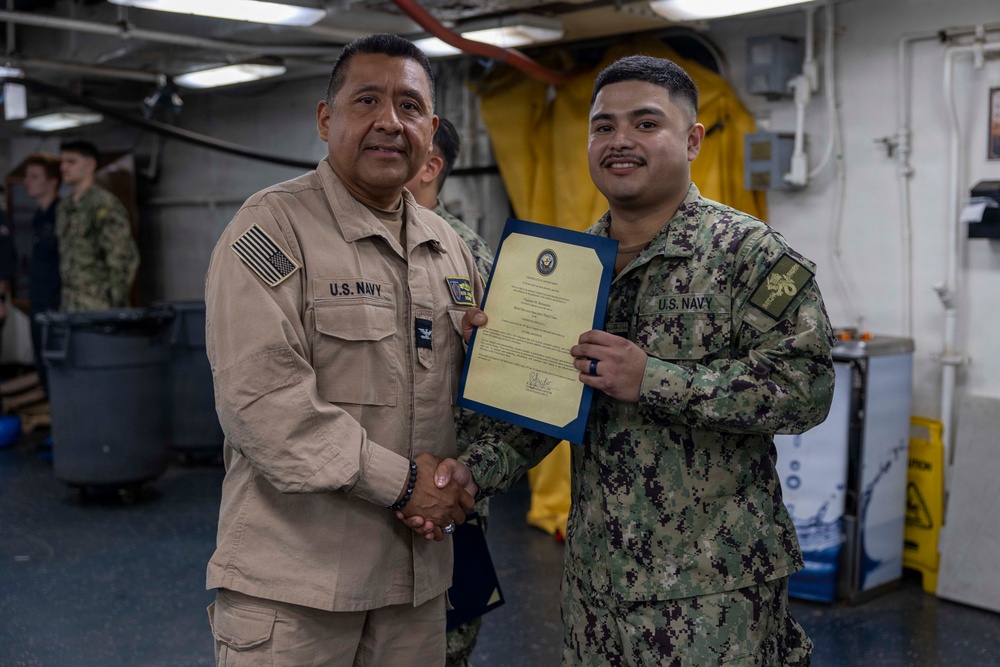 Two U.S. Navy personnel are shaking hands during what appears to be a formal recognition or award ceremony aboard a naval vessel. The senior officer on the left, wearing khaki service uniform with a captain's eagle insignia, is presenting a framed certificate to a junior sailor in Navy Working Uniform (NWU Type III digital camouflage). Both individuals are smiling, suggesting a positive ceremonial occasion.
