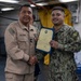 Two U.S. Navy personnel are shaking hands during what appears to be a formal recognition or award ceremony aboard a naval vessel. The senior officer on the left, wearing khaki service uniform with a captain's eagle insignia, is presenting a framed certificate to a junior sailor in Navy Working Uniform (NWU Type III digital camouflage). Both individuals are smiling, suggesting a positive ceremonial occasion.