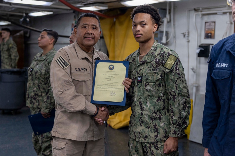 Two U.S. Navy personnel are shaking hands while jointly holding a framed certificate or commendation document aboard what appears to be a naval vessel. The senior sailor in tan/khaki uniform is presenting the award to a junior sailor in Navy Working Uniform (NWU) camouflage pattern. Additional Navy personnel are visible in the background observing the ceremony.
