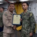 Two U.S. Navy personnel are shaking hands while jointly holding a framed certificate or commendation document aboard what appears to be a naval vessel. The senior sailor in tan/khaki uniform is presenting the award to a junior sailor in Navy Working Uniform (NWU) camouflage pattern. Additional Navy personnel are visible in the background observing the ceremony.
