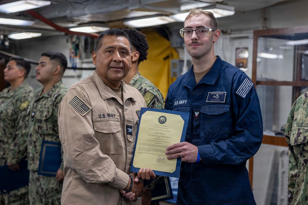 Two U.S. Navy personnel are shaking hands while jointly holding a certificate or commendation document during what appears to be an awards ceremony aboard a naval vessel. The senior sailor on the left, wearing a tan/khaki uniform, is presenting the document to a junior sailor on the right wearing a blue coverall uniform. Several other Navy personnel in camouflage uniforms are visible in the background.