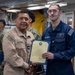 Two U.S. Navy personnel are shaking hands while jointly holding a certificate or commendation document during what appears to be an awards ceremony aboard a naval vessel. The senior sailor on the left, wearing a tan/khaki uniform, is presenting the document to a junior sailor on the right wearing a blue coverall uniform. Several other Navy personnel in camouflage uniforms are visible in the background.