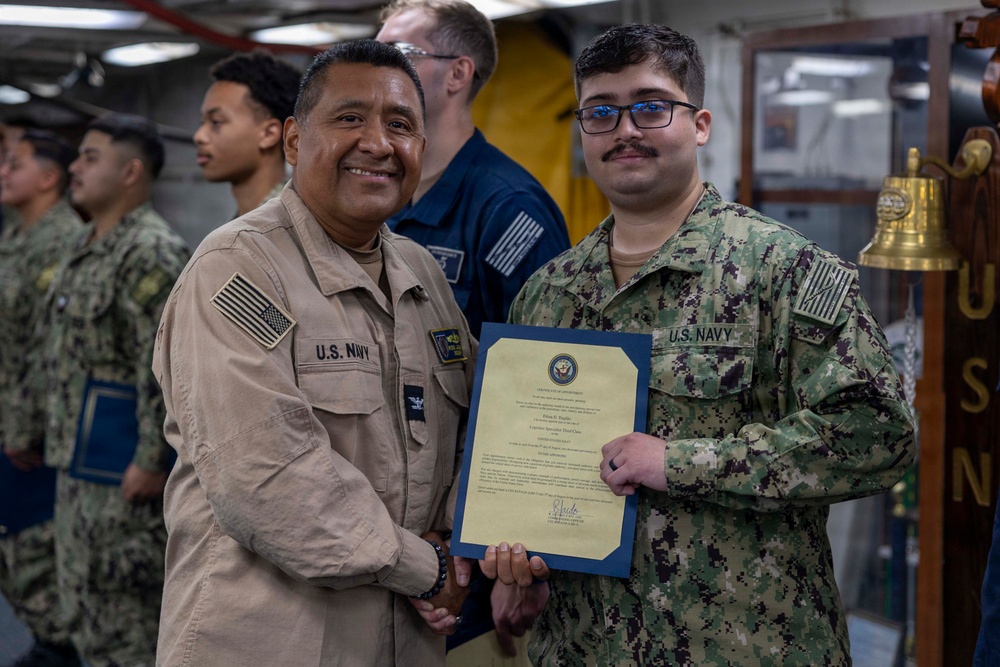 Two U.S. Navy personnel are posing together while jointly holding a certificate or commendation document with a blue folder and yellow paper. The ceremony appears to be taking place aboard a naval vessel, with other sailors visible in the background. One sailor wears khaki service uniform while the other wears the Navy Working Uniform (NWU) digital camouflage pattern.