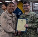 Two U.S. Navy personnel are posing together while jointly holding a certificate or commendation document with a blue folder and yellow paper. The ceremony appears to be taking place aboard a naval vessel, with other sailors visible in the background. One sailor wears khaki service uniform while the other wears the Navy Working Uniform (NWU) digital camouflage pattern.