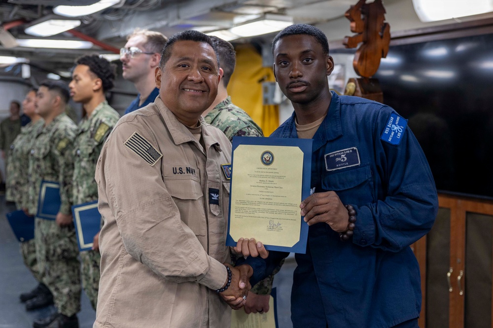 A U.S. Navy officer in tan/khaki uniform is presenting a certificate or commendation document to a junior sailor in blue coveralls aboard what appears to be a naval vessel. The two individuals are shaking hands while jointly holding the framed certificate. Additional sailors in camouflage uniforms are visible in the background, suggesting a formal recognition ceremony.