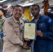 A U.S. Navy officer in tan/khaki uniform is presenting a certificate or commendation document to a junior sailor in blue coveralls aboard what appears to be a naval vessel. The two individuals are shaking hands while jointly holding the framed certificate. Additional sailors in camouflage uniforms are visible in the background, suggesting a formal recognition ceremony.