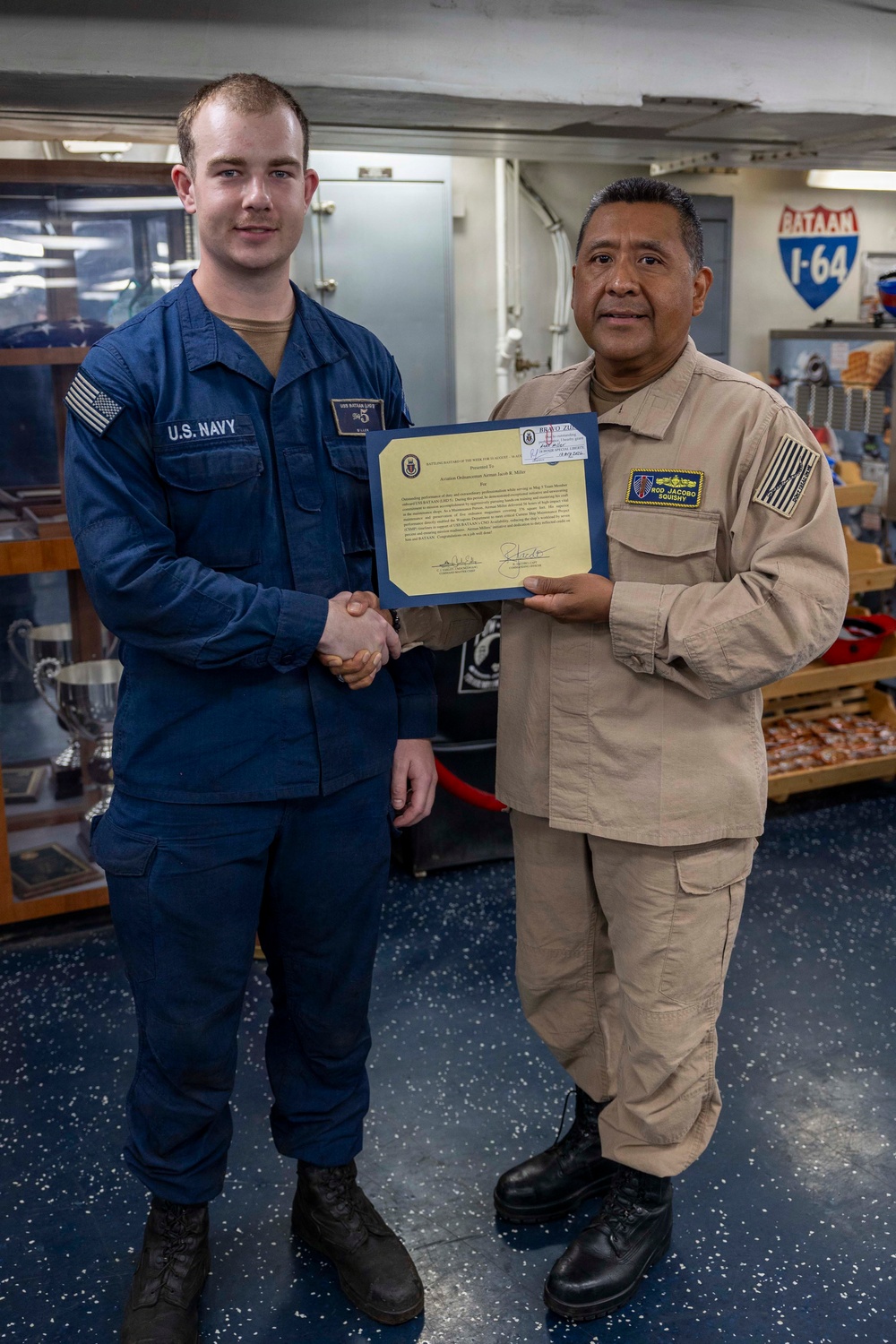 Two U.S. Navy personnel are posing for a photo aboard what appears to be USS Bataan (LHD-5), with one sailor in blue Navy working uniform and one in tan/khaki uniform shaking hands while jointly holding a certificate or award. The ceremony appears to be a recognition event, likely a 'Sailor of the Week' or similar commendation presentation. The setting is an interior space of a naval vessel with trophies and ship insignia visible in the background.