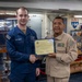 Two U.S. Navy personnel are posing for a photo aboard what appears to be USS Bataan (LHD-5), with one sailor in blue Navy working uniform and one in tan/khaki uniform shaking hands while jointly holding a certificate or award. The ceremony appears to be a recognition event, likely a 'Sailor of the Week' or similar commendation presentation. The setting is an interior space of a naval vessel with trophies and ship insignia visible in the background.